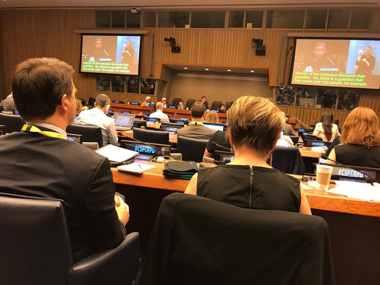 Attendees facing a United Nations session with live sign language interpretation shown on large screens